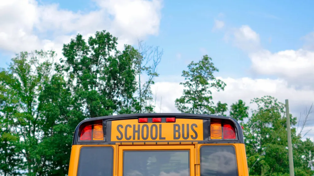 The SCHOOL BUS sign and warning lights on a yellow bus, framed by green treetops and a blue sky with white clouds