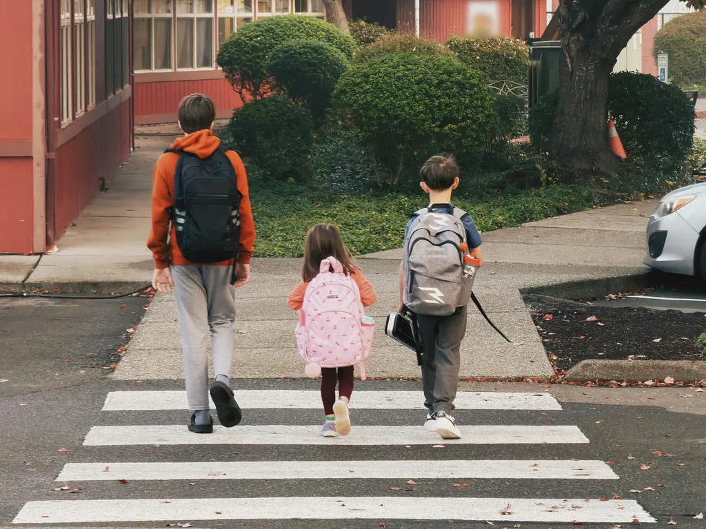 Three children wearing backpacks walking across a crosswalk, seen from behind